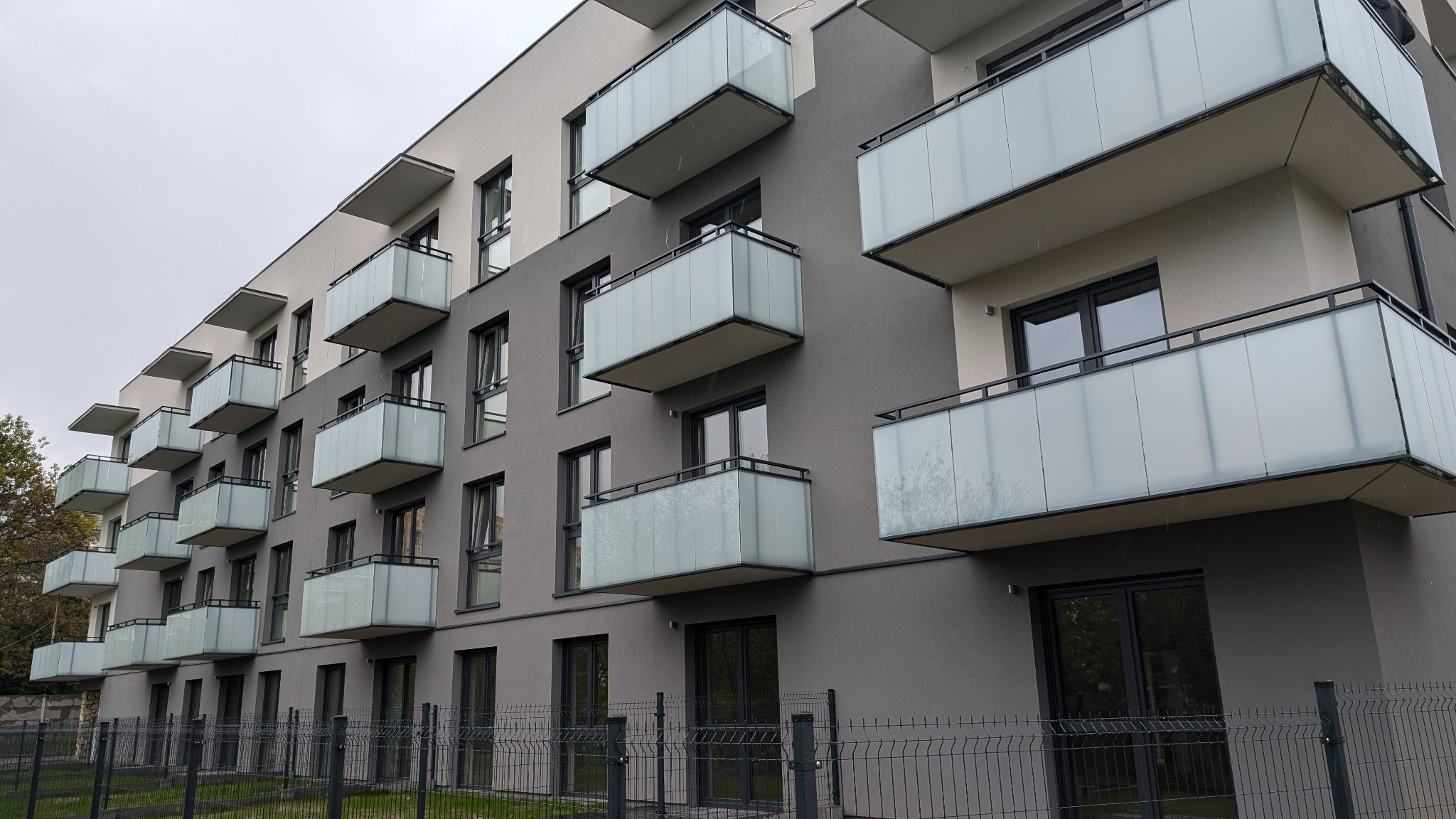 Nowoczesny budynek z balkonami o szklanych balustradach, czarne ramy okienne i subtelna kolorystyka elewacji w odcieniach szarości i bieli. Minimalistyczny design.