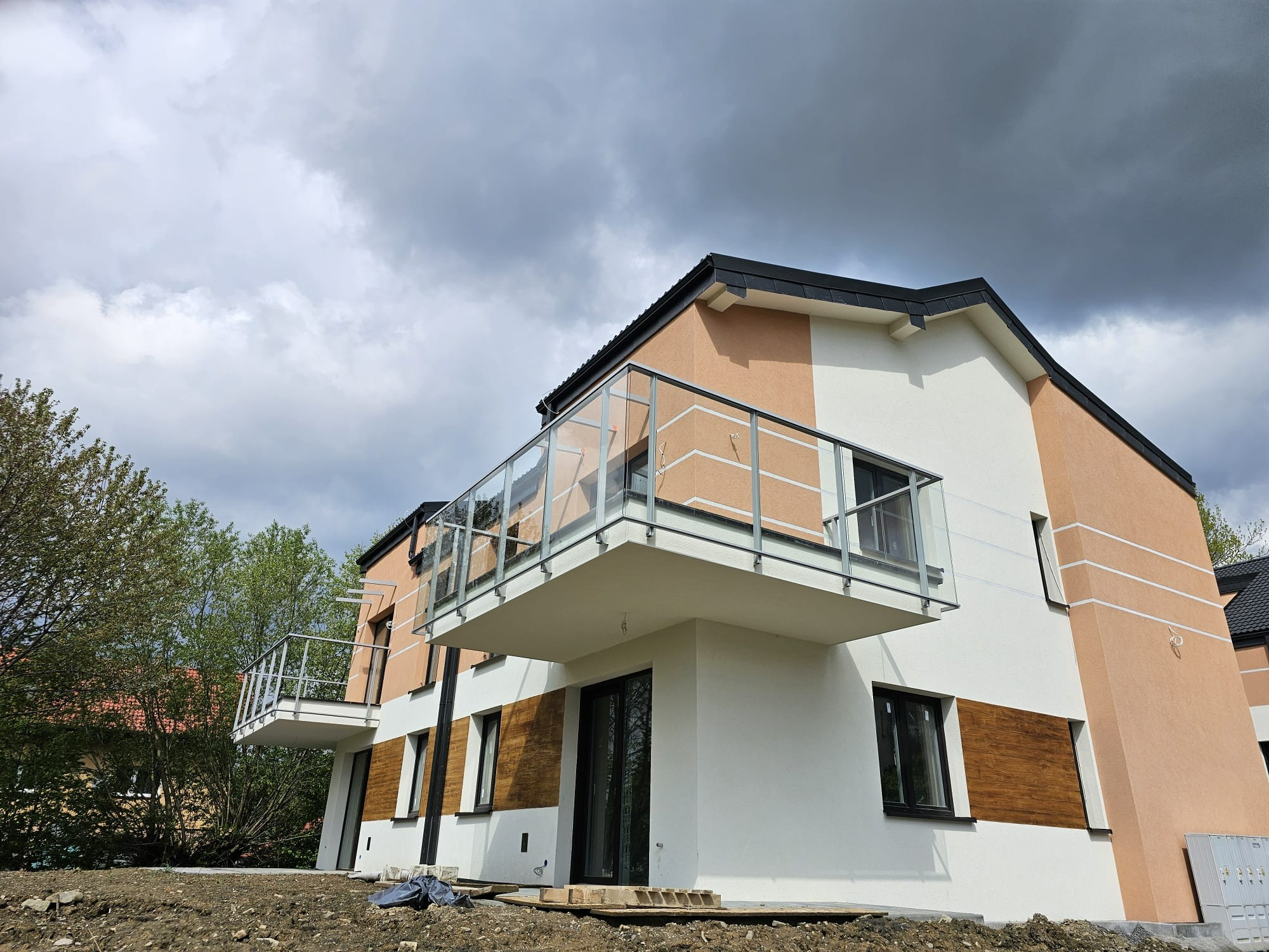 Nowoczesny budynek z balkonami z balustradami szklanymi i metalowymi, widok z dołu, pochmurne niebo w tle. Budynek w kolorach bieli i beżu.