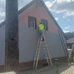 Złota rączka - Pracownik na drabinie maluje elewację domu na szaro, z pomarańczowymi akcentami wokół okien. Częściowo pomalowana ściana, widoczne dwie drabiny.