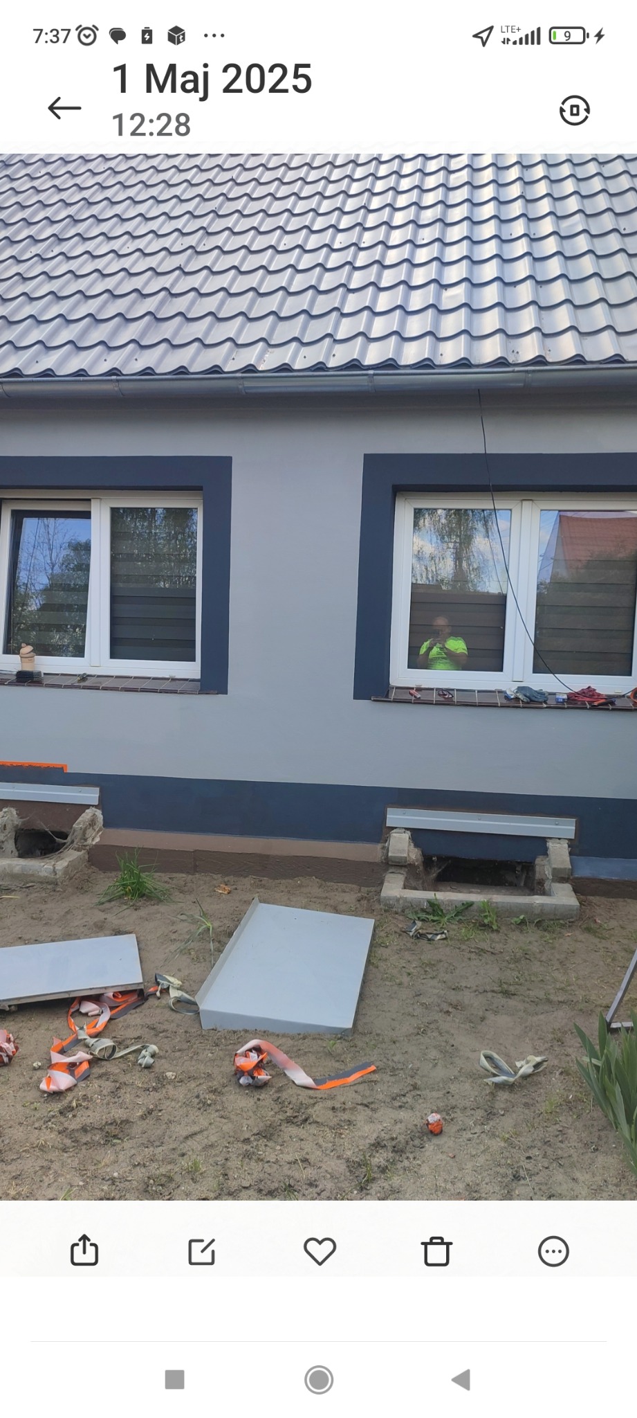 Szara elewacja domu z dwoma oknami, widoczne elementy montażowe i gruz przed budynkiem, sugerujące prace wykończeniowe lub remontowe.