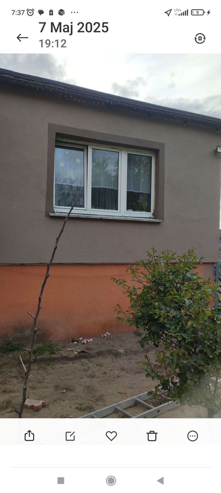 Dom z oknem po remoncie elewacji, widoczny fragment drabiny i krzew. Świeżo pomalowana fasada w odcieniach beżu i pomarańczy. Nowoczesne okno z białymi ramami.