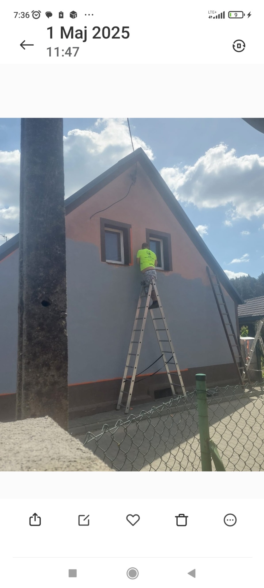 Pracownik na drabinie maluje elewację domu na szaro, z pomarańczowymi akcentami wokół okien. Częściowo pomalowana ściana, widoczne dwie drabiny.