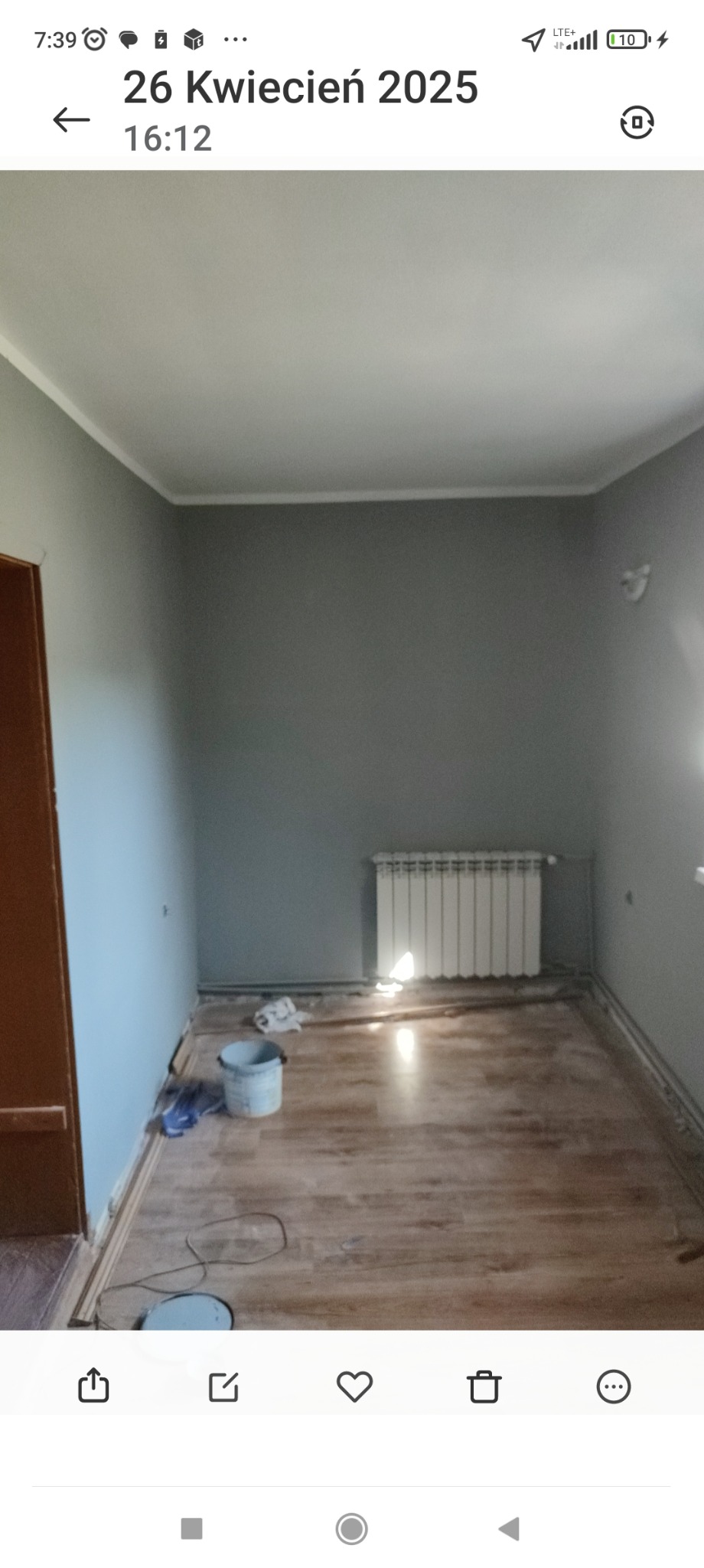 Pomieszczenie po malowaniu ścian na szaro, widoczny grzejnik, podłoga i wiadro z farbą. Wykończenia wnętrz w trakcie prac.