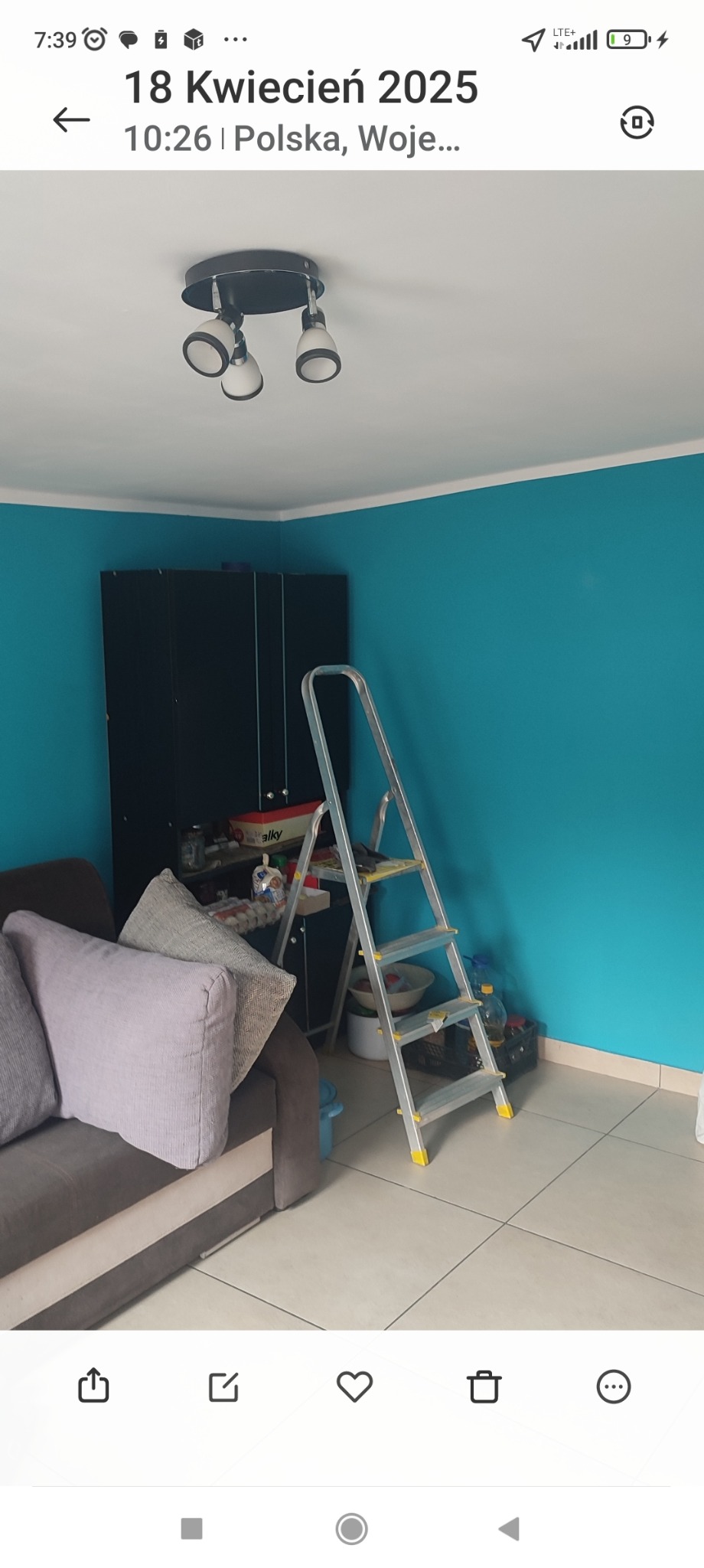 Ściana pomalowana na turkusowo, drabina aluminiowa, czarna szafa w rogu pokoju. Widoczne również oświetlenie sufitowe i fragment sofy z poduszkami.