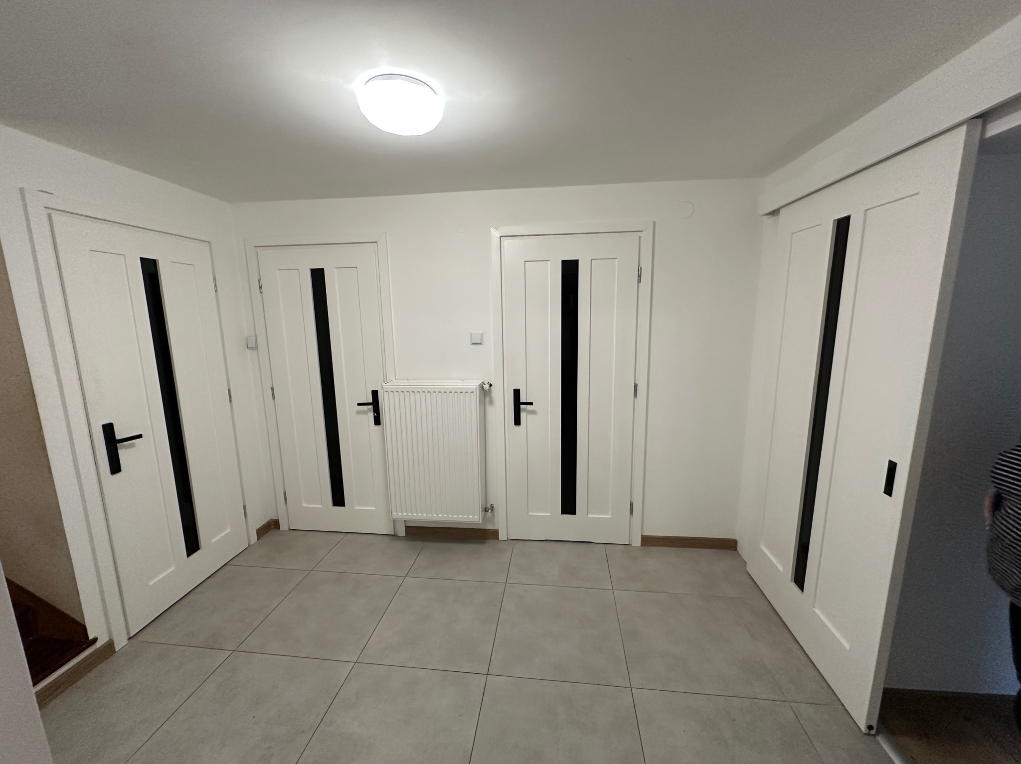 Nowoczesny przedpokój z trzema białymi, drewnianymi drzwiami z czarnymi wstawkami i klamkami, szarą podłogą z płytek i białym grzejnikiem.