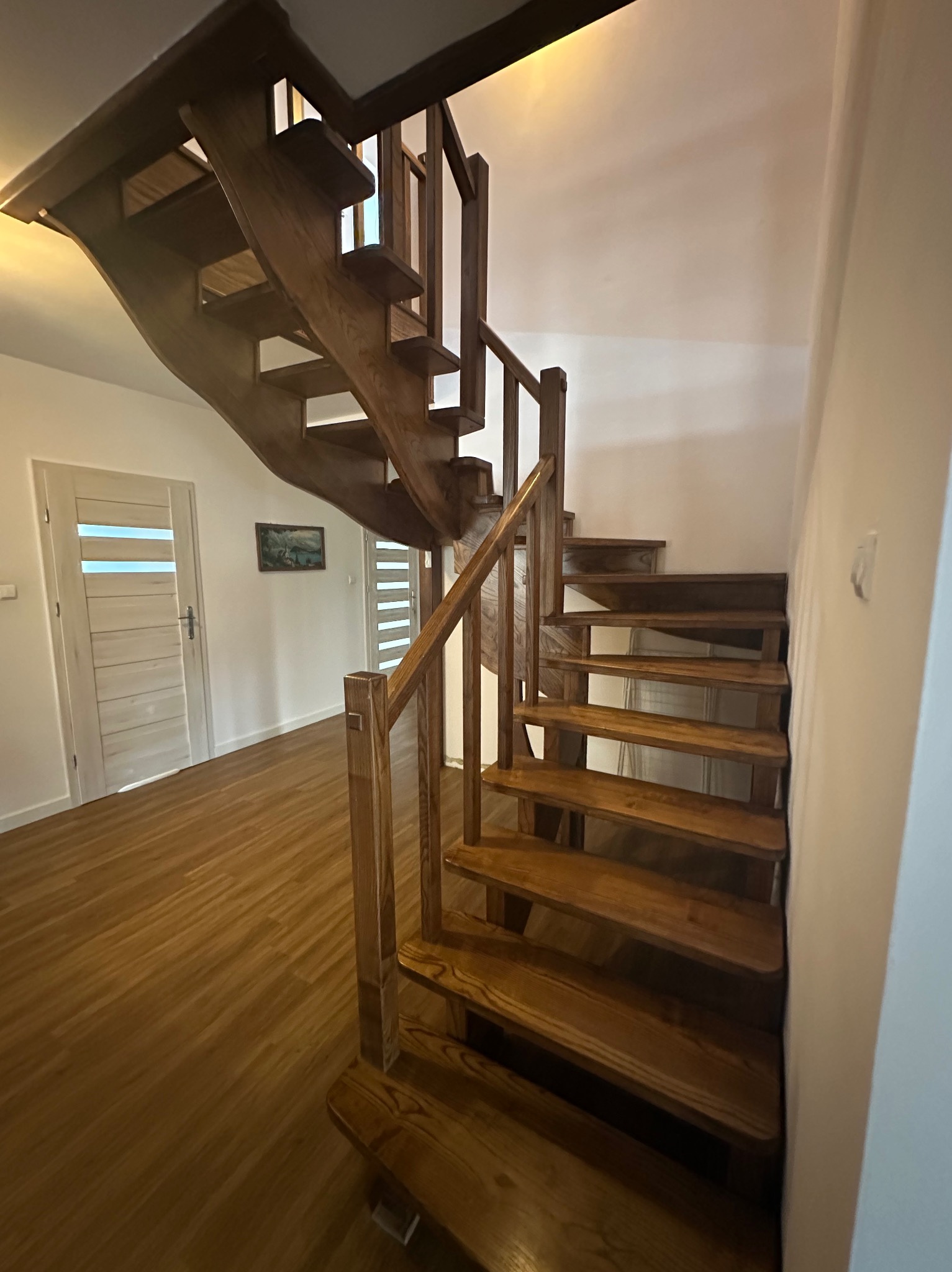 Drewniane schody z balustradą w jasnym wnętrzu. Widoczne detale konstrukcyjne, naturalne oświetlenie i wykończenie podłogi. Dwie pary drzwi w tle.