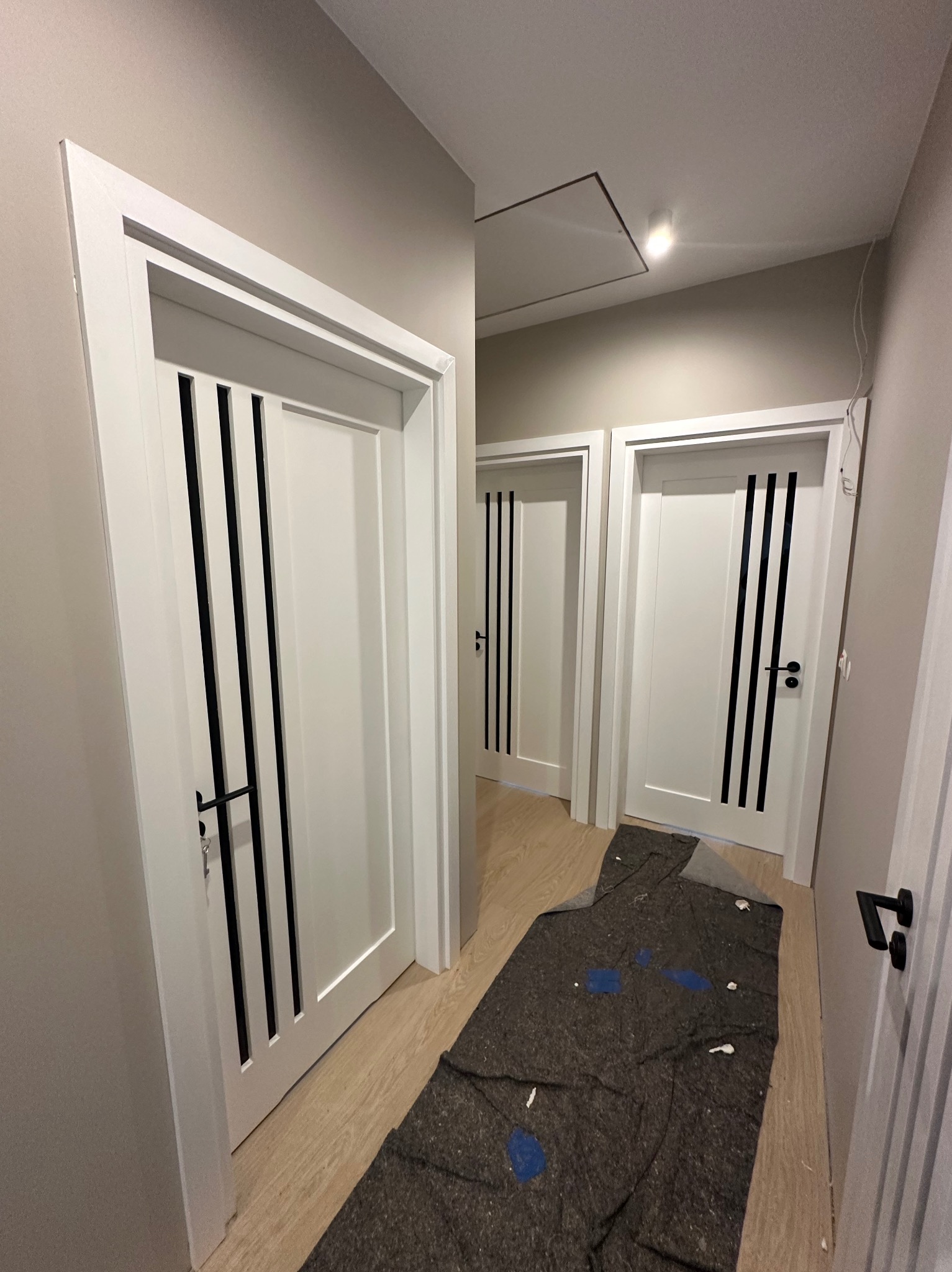 Białe drzwi wewnętrzne z czarnymi, pionowymi zdobieniami w korytarzu. Ochronna mata budowlana na podłodze. Widoczne trzy pary drzwi w perspektywie.