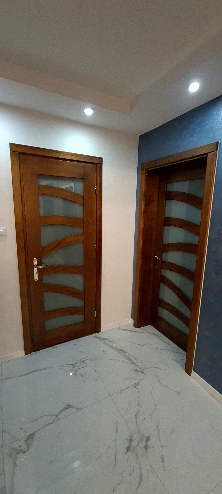 Dwa drewniane drzwi z przeszkleniami o falistym wzorze. Jedne drzwi na białej ścianie, drugie na ścianie w odcieniach granatu. Marmurowe płytki podłogowe.