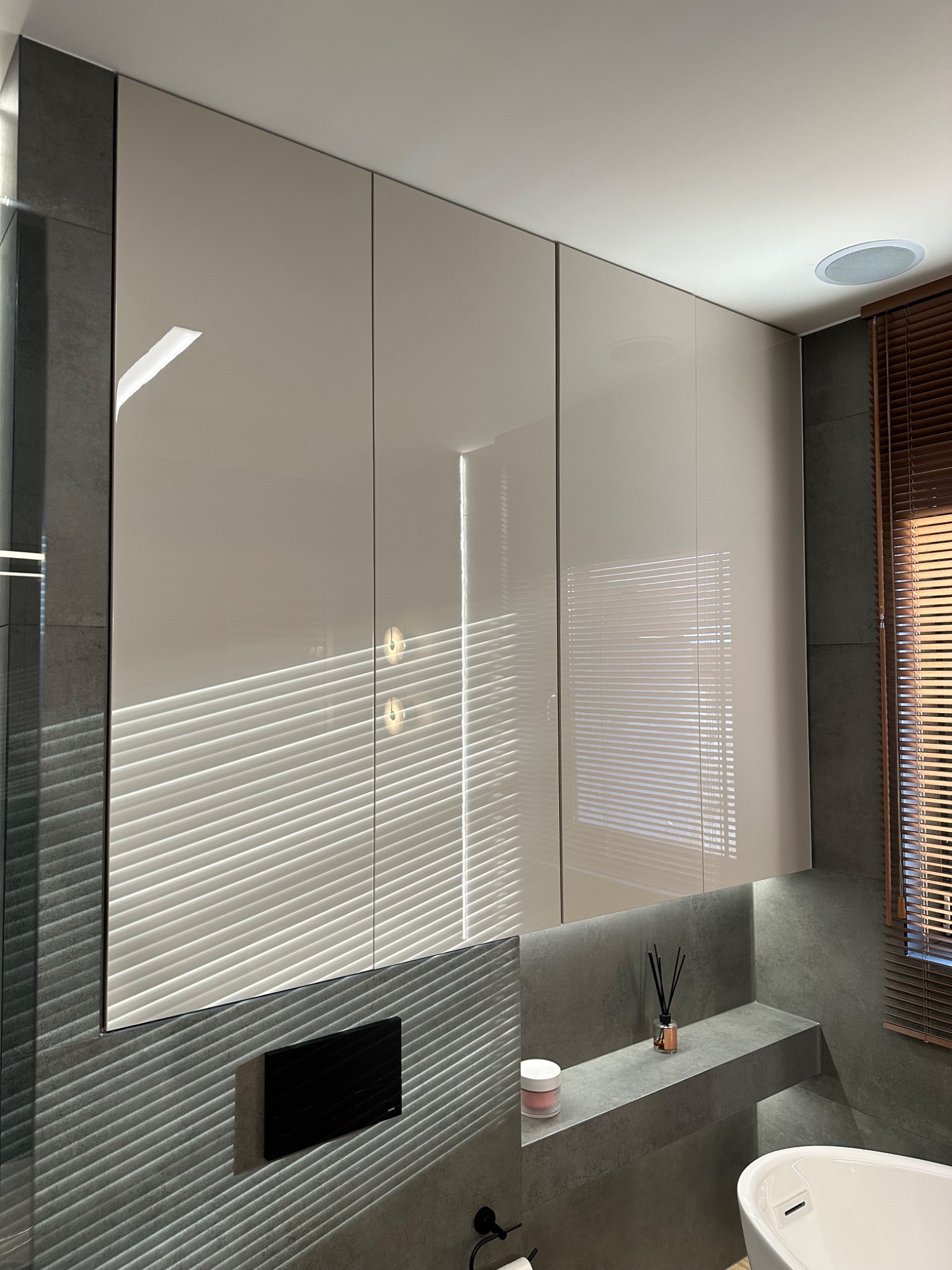 Beżowe szafki łazienkowe pod sufitem, z odbiciem żaluzji. Szara ściana z półką i dekoracjami. Minimalistyczny design, nowoczesne wykończenie.