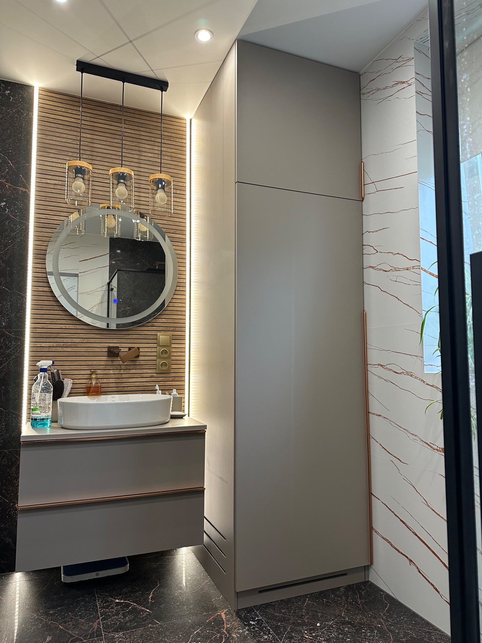 Nowoczesna łazienka z szarą szafką pod umywalką, okrągłym lustrem z podświetleniem LED i wysoką szafą w zabudowie. Uwagę zwraca drewniana ściana i marmurowe płytki.