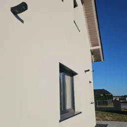 Jasna elewacja domu jednorodzinnego z widocznymi oknami z czarną ramą, lampą zewnętrzną i fragmentem drewnianej podbitki dachowej, obok wyłożony kamieniami ozdobnymi pas i chodnik z kostki brukowej.