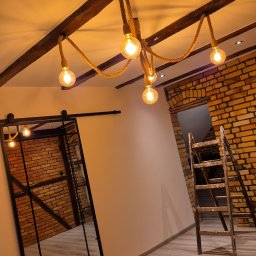 Budomex Sebastian Ślusarczyk - Loftowe wnętrze z ceglaną ścianą, drewnianymi belkami sufitowymi i lampą z żarówek na sznurze. Przesuwne drzwi z czarnymi szprosami odbijają ceglany mur. Drewniana drabina oparta o ścianę.
