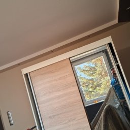 Budomex Sebastian Ślusarczyk - Wnętrze pokoju w trakcie prac wykończeniowych, widoczna ściana pomalowana na dwa odcienie beżu i szarości, przesuwana szafa z okleiną imitującą drewno i okno z widokiem na drzewo, częściowo zakryte...