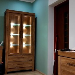 Budomex Sebastian Ślusarczyk - Wnętrze pokoju z drewnianą witryną z podświetlanymi półkami, widoczna miętowa ściana i drewniana podłoga, obok stoi komoda i oparta o ścianę zmiotka.