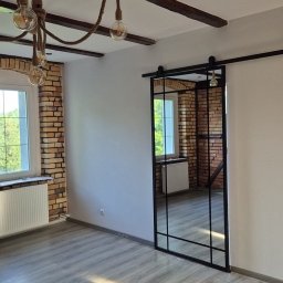 Budomex Sebastian Ślusarczyk - Jasne, przestronne wnętrze z ceglaną ścianą, drewnianymi belkami stropowymi i lampami na sznurze, z przesuwnymi drzwiami w stylu loft z czarnymi szprosami, odbijającymi fragment pokoju.