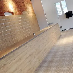 Surowe wnętrze sklepu z elementami cegły, drewnianą ladą z metalowymi listwami i wzorzystą podłogą, oświetlone reflektorami na belkach stropowych.