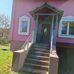 Budomex Sebastian Ślusarczyk - Różowa elewacja domu z szarym zadaszeniem nad wejściem i betonowymi schodami z metalową barierką.