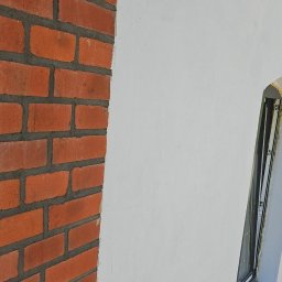 Budomex Sebastian Ślusarczyk - Fragment elewacji budynku z ceglaną ścianą i oknem obok otynkowanej powierzchni, widoczne ślady po szpachlowaniu.