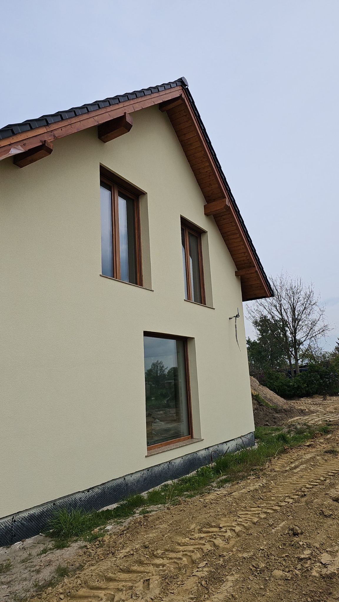Fragment nowo wybudowanego domu z beżową elewacją, drewnianymi oknami i czarną dachówką, widoczne ślady po budowie na działce, pochmurne niebo.