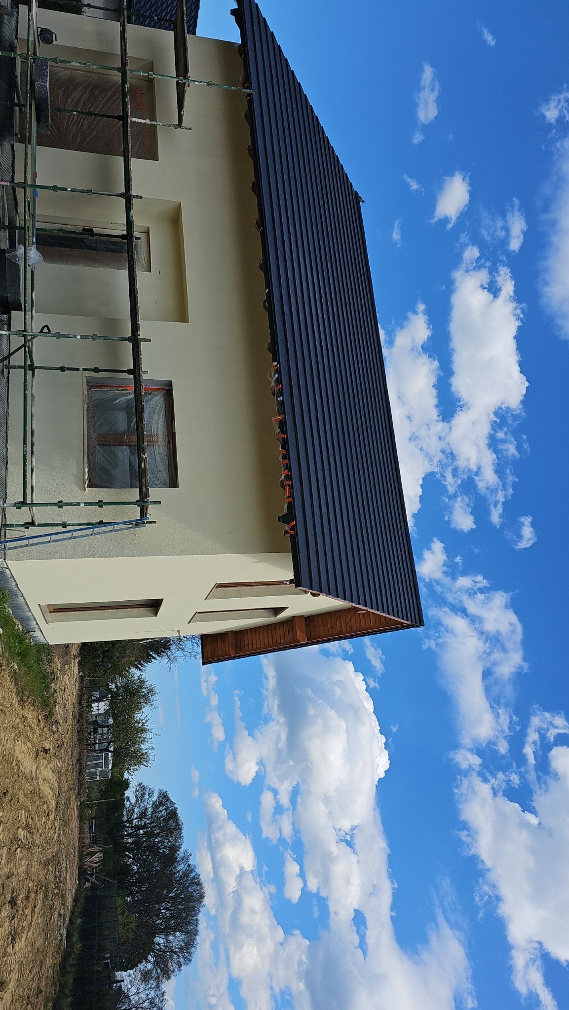 Dom jednorodzinny w trakcie prac elewacyjnych, widoczne rusztowania, zabezpieczone okna folią, fragment nowej, ciemnej elewacji, błękitne niebo z chmurami w tle.