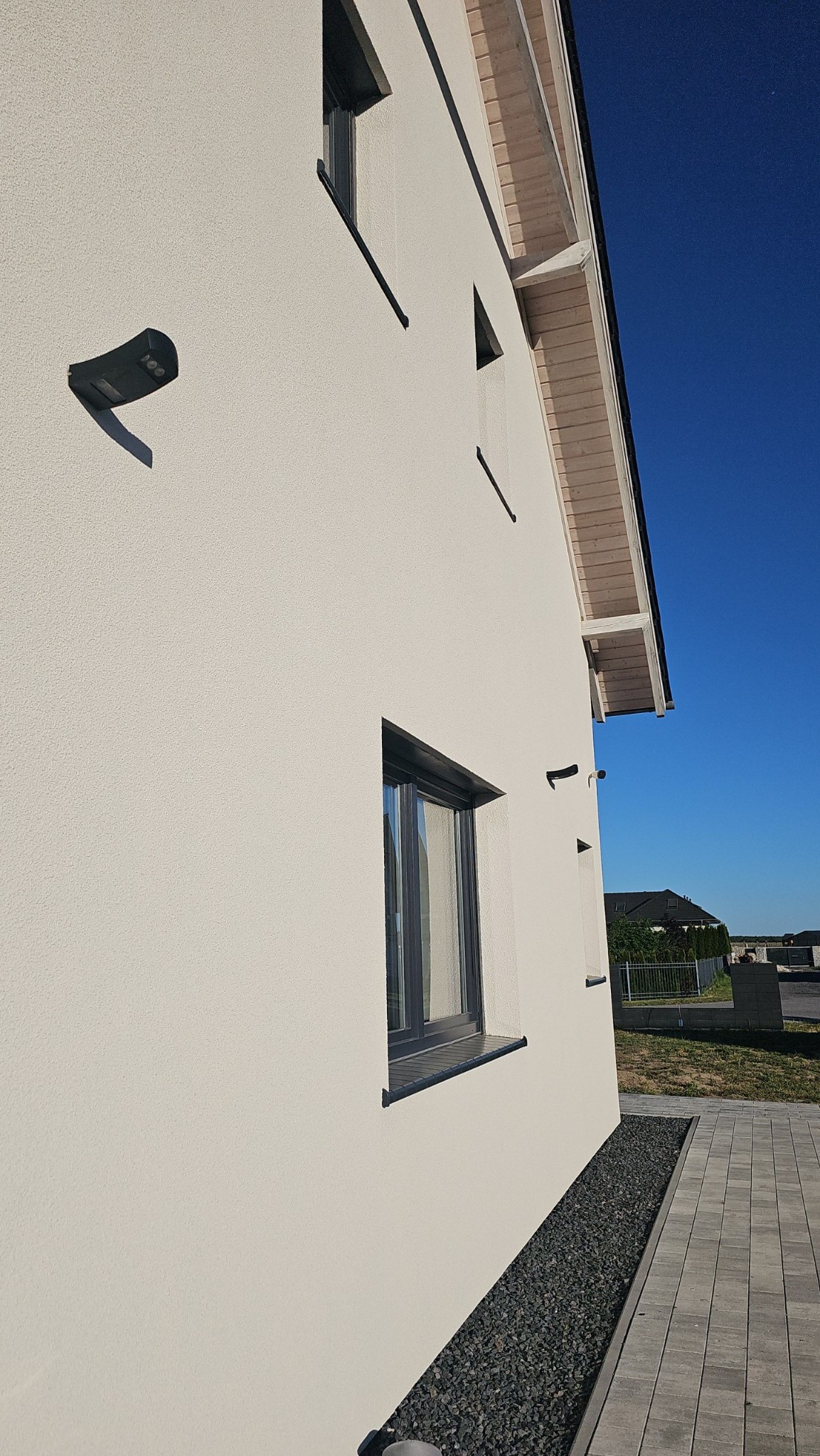 Jasna elewacja domu jednorodzinnego z widocznymi oknami z czarną ramą, lampą zewnętrzną i fragmentem drewnianej podbitki dachowej, obok wyłożony kamieniami ozdobnymi pas i chodnik z kostki brukowej.