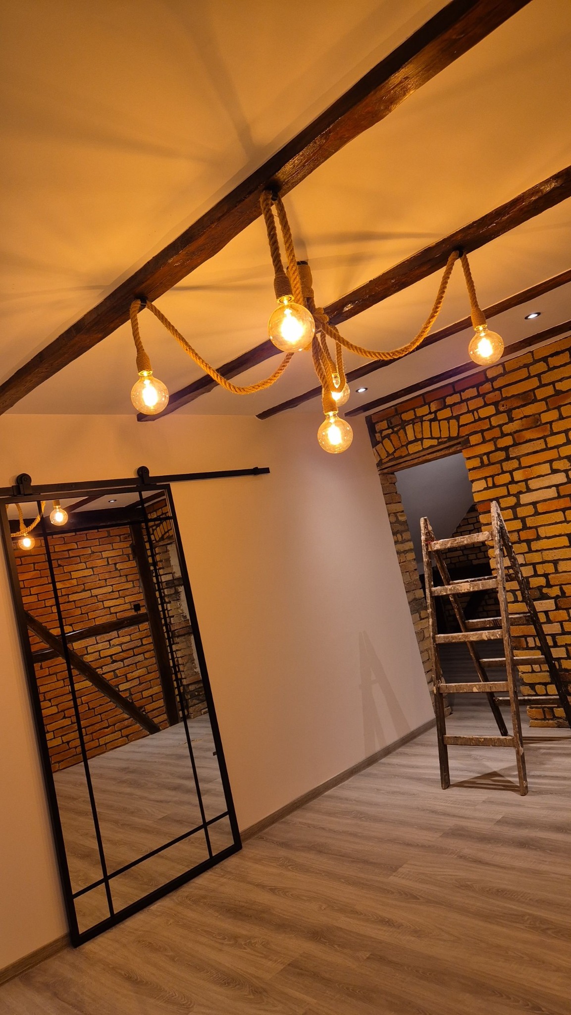 Loftowe wnętrze z ceglaną ścianą, drewnianymi belkami sufitowymi i lampą z żarówek na sznurze. Przesuwne drzwi z czarnymi szprosami odbijają ceglany mur. Drewniana drabina oparta o ścianę.
