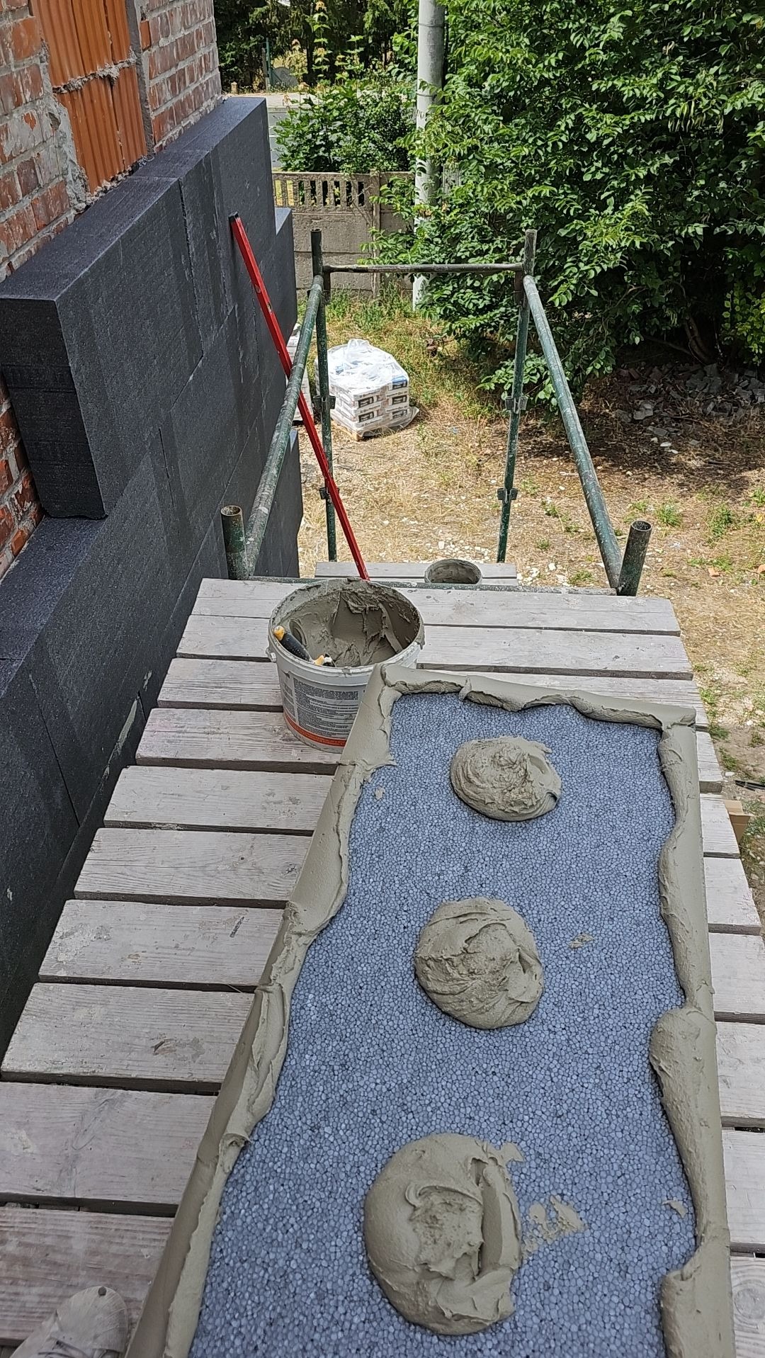 Przygotowanie płyty styropianowej z nałożonym klejem w formie placków i obrzeży, widoczny fragment ściany z przyklejonymi płytami, wiadro z klejem i poziomica na rusztowaniu.