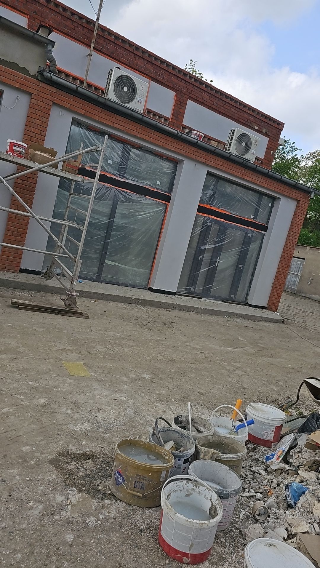 Budynek w trakcie prac elewacyjnych: ceglano-szare wykończenie, okna zabezpieczone folią, widoczne rusztowanie i wiaderka z zaprawą na ziemi.