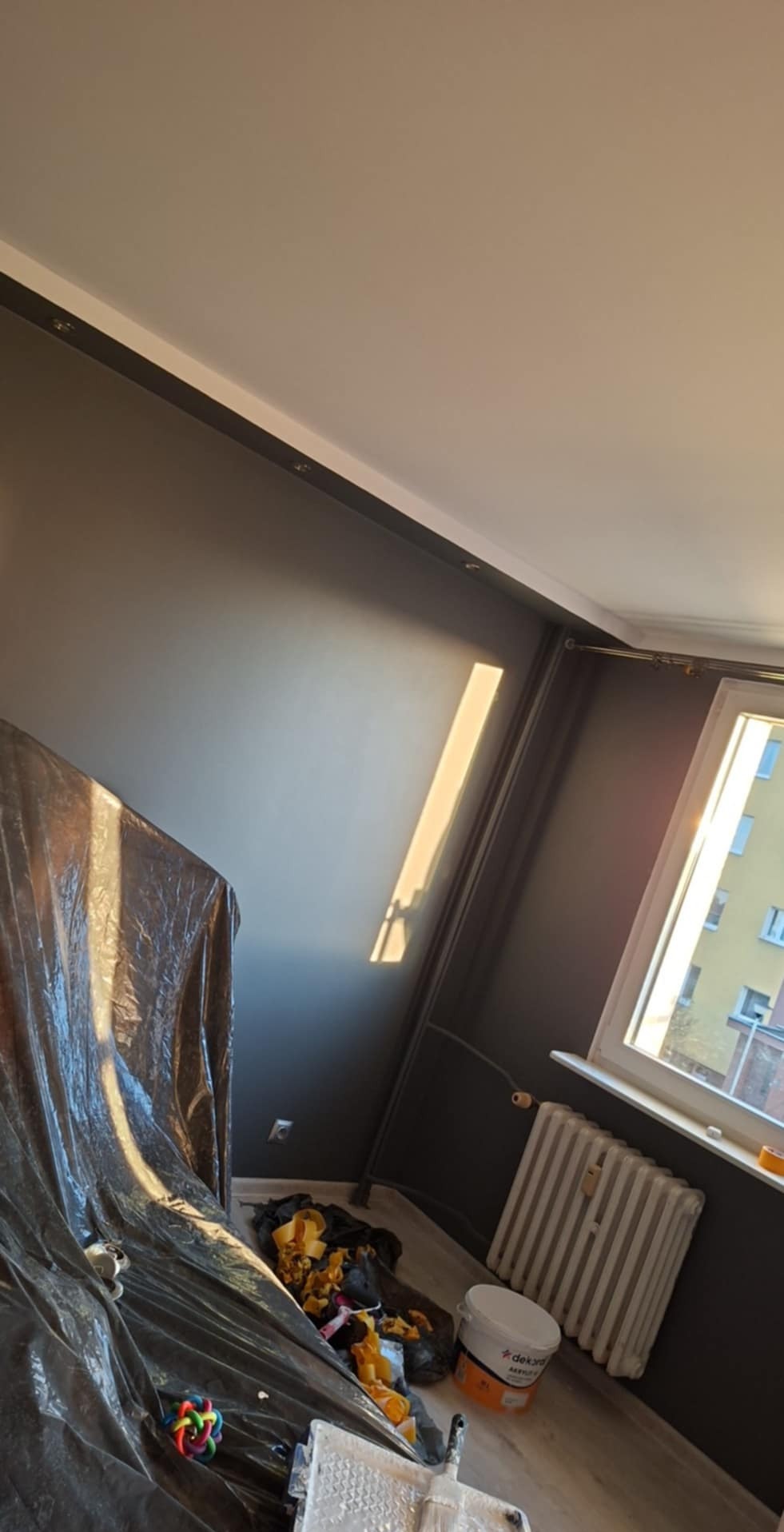 Pokój w trakcie malowania na ciemny kolor, zabezpieczony folią, widoczny grzejnik, puszka z farbą Dekoral i narzędzia malarskie w nieładzie.