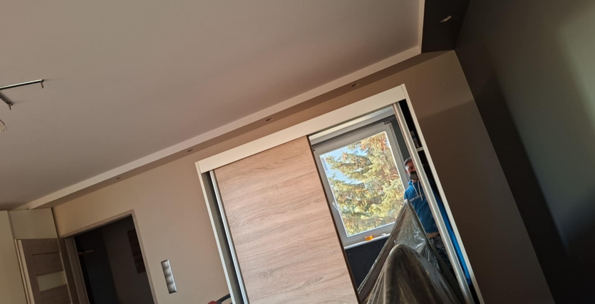 Wnętrze pokoju w trakcie prac wykończeniowych, widoczna ściana pomalowana na dwa odcienie beżu i szarości, przesuwana szafa z okleiną imitującą drewno i okno z widokiem na drzewo, częściowo zakryte...