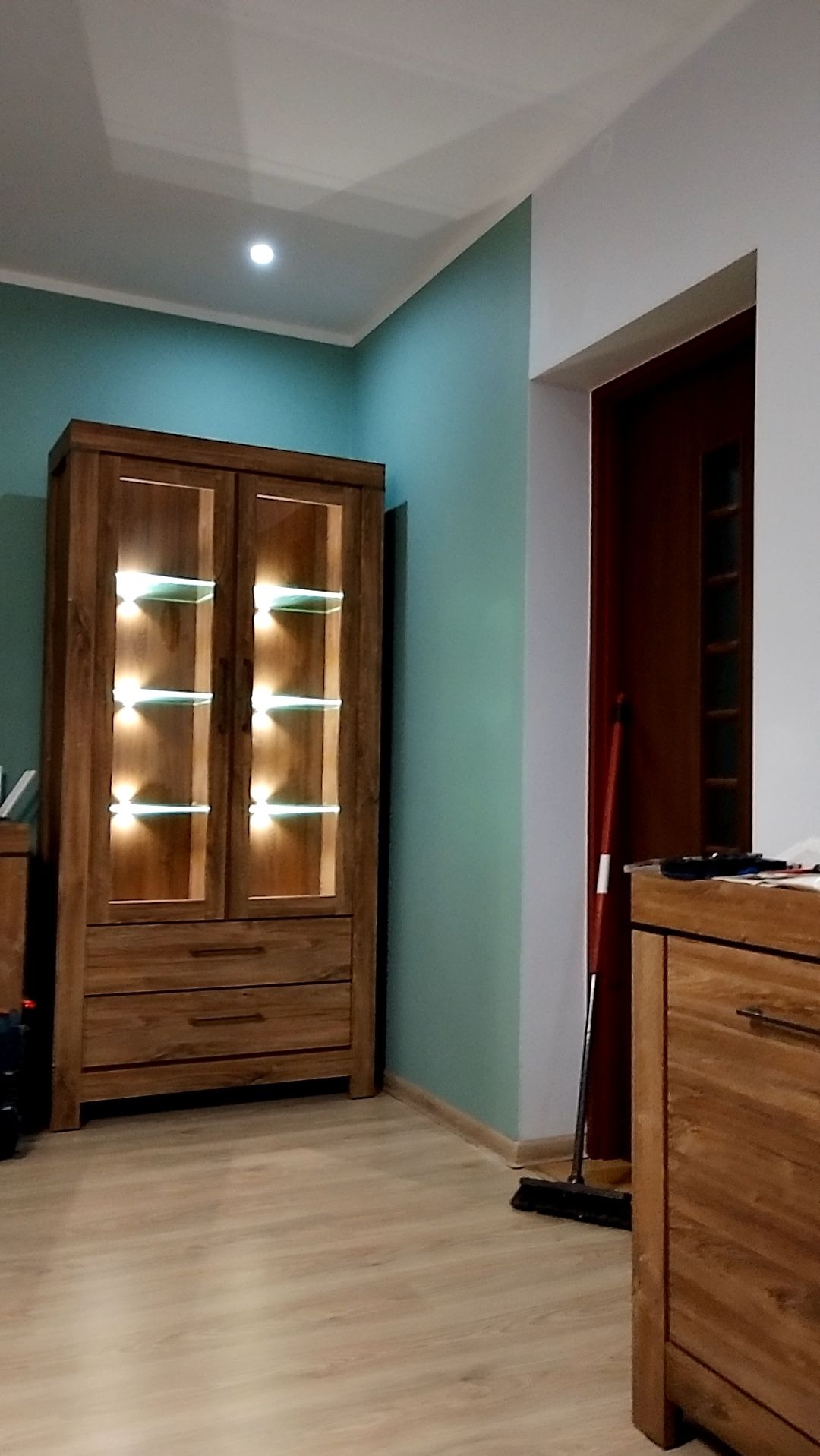 Wnętrze pokoju z drewnianą witryną z podświetlanymi półkami, widoczna miętowa ściana i drewniana podłoga, obok stoi komoda i oparta o ścianę zmiotka.