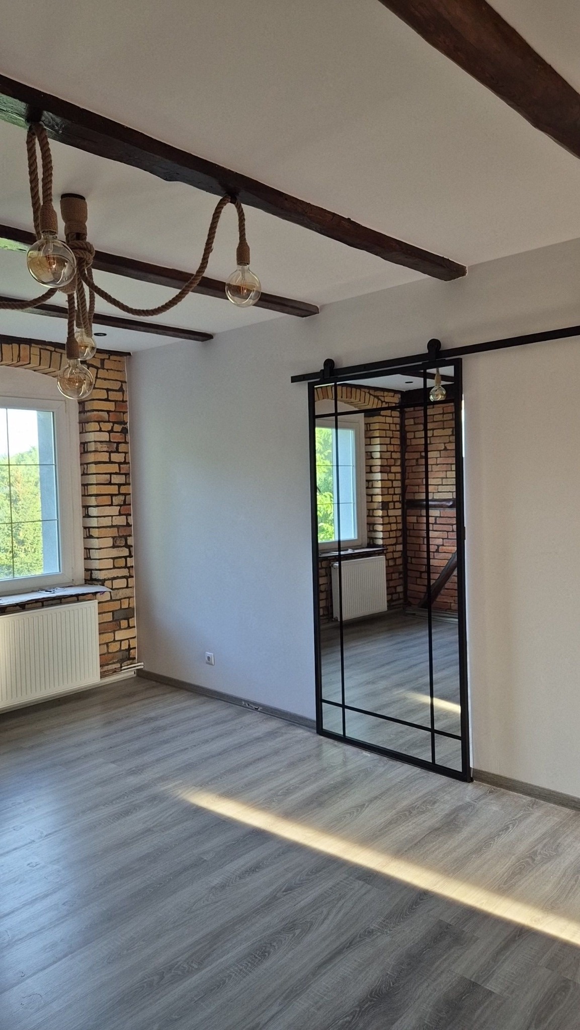 Jasne, przestronne wnętrze z ceglaną ścianą, drewnianymi belkami stropowymi i lampami na sznurze, z przesuwnymi drzwiami w stylu loft z czarnymi szprosami, odbijającymi fragment pokoju.