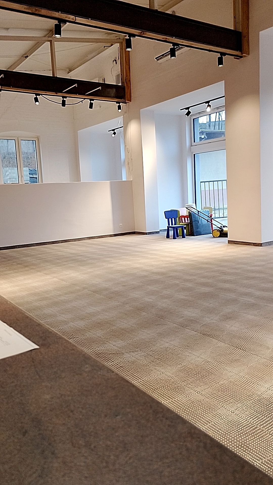 Przestronne, jasne wnętrze biurowe z beżową wykładziną w drobną kratkę, surowymi belkami stropowymi z oświetleniem punktowym i widokiem na zewnątrz przez duże okno z niebieskim krzesełkiem...