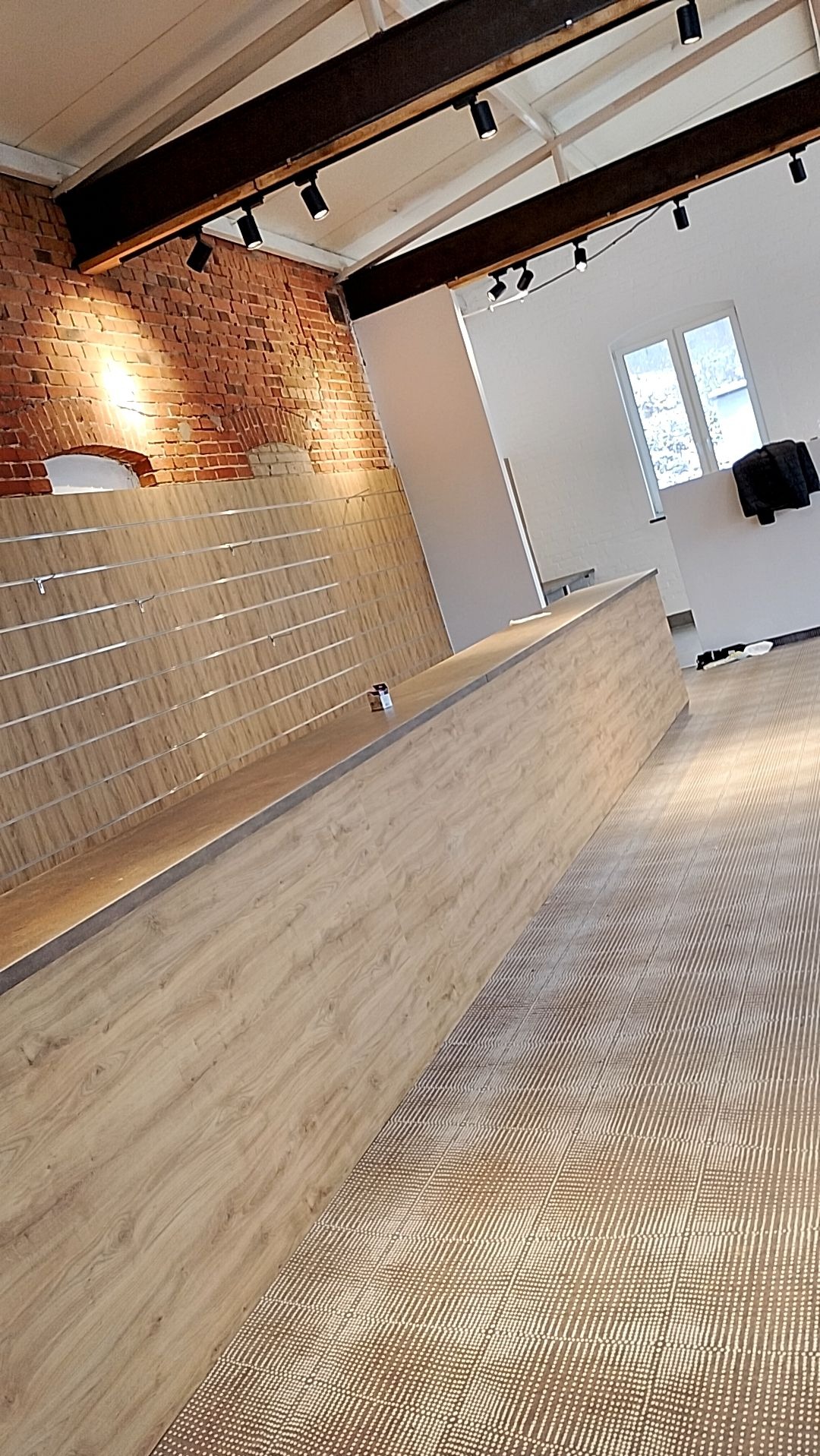 Surowe wnętrze sklepu z elementami cegły, drewnianą ladą z metalowymi listwami i wzorzystą podłogą, oświetlone reflektorami na belkach stropowych.