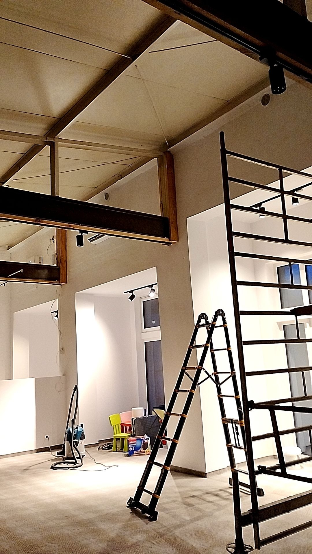 Wnętrze remontowanego pomieszczenia z wysokim sufitem, widoczne metalowe belki konstrukcyjne, drabina, odkurzacz przemysłowy i meble w tle.