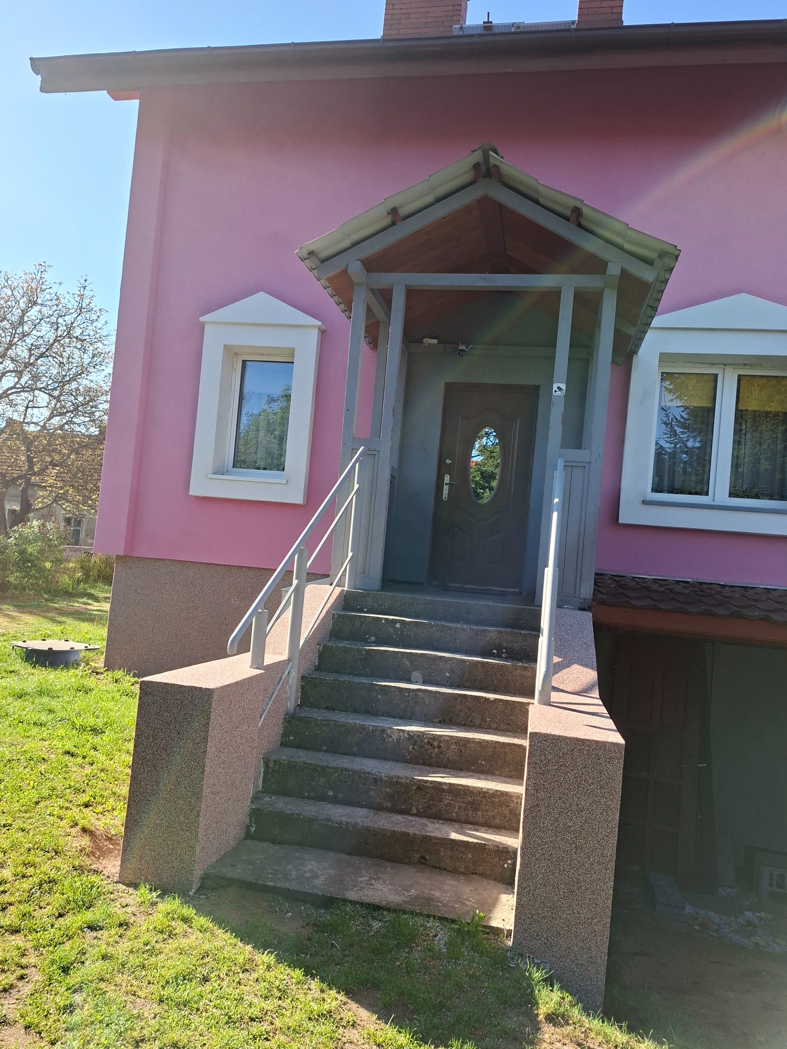Różowa elewacja domu z szarym zadaszeniem nad wejściem i betonowymi schodami z metalową barierką.