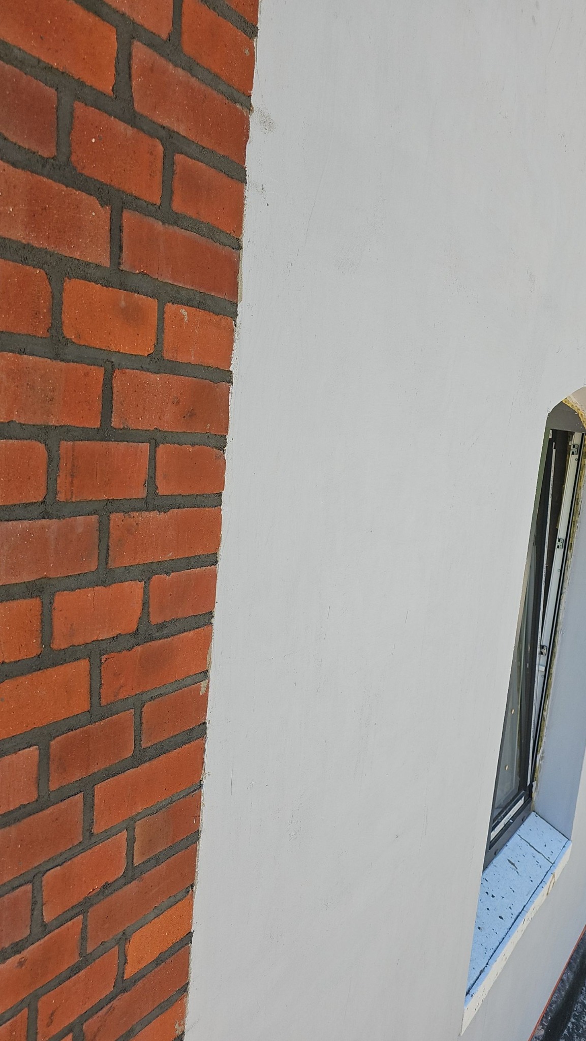 Fragment elewacji budynku z ceglaną ścianą i oknem obok otynkowanej powierzchni, widoczne ślady po szpachlowaniu.