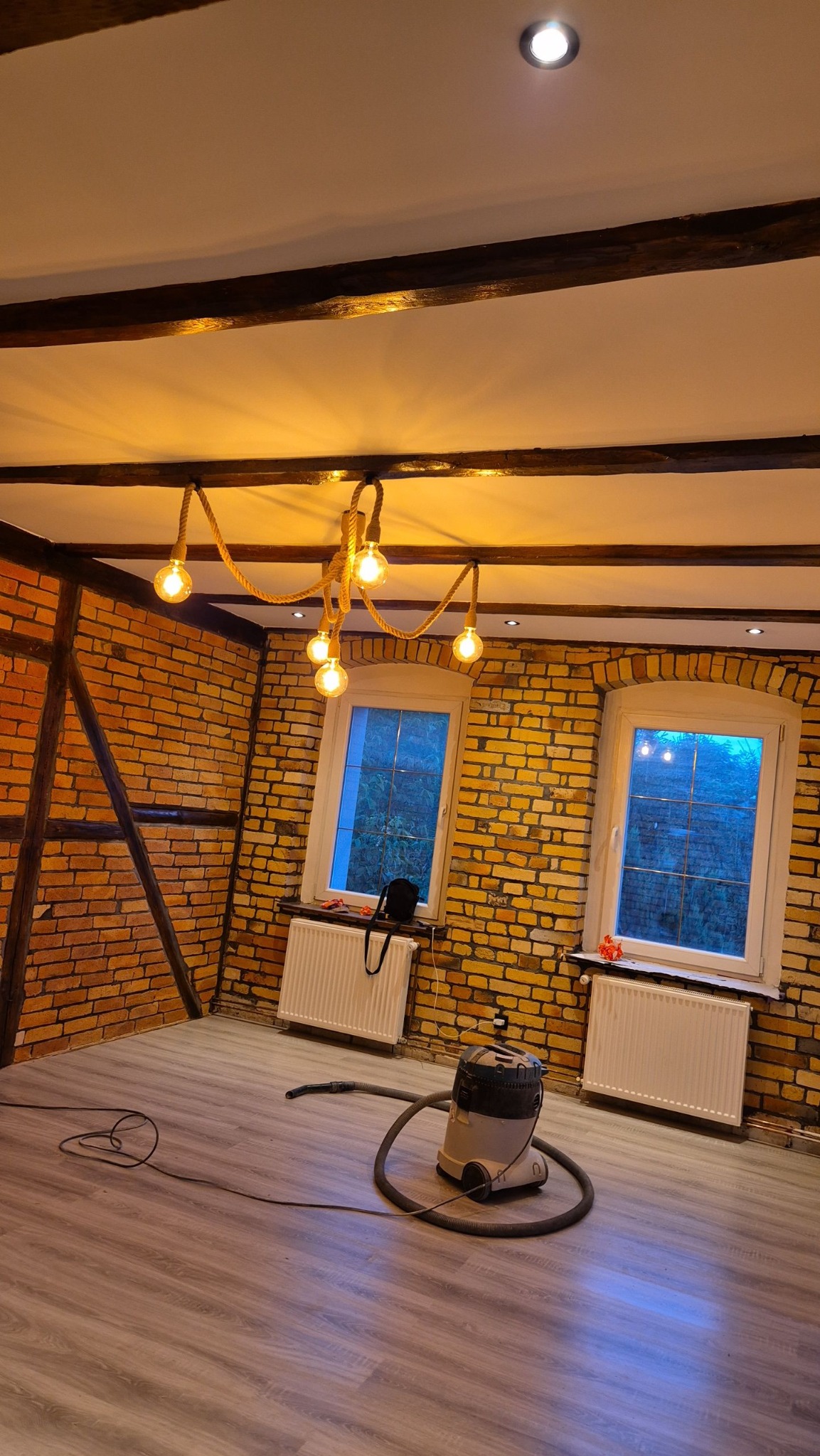 Pokój po remoncie z odsłoniętą cegłą na ścianach, drewnianymi belkami pod sufitem i oryginalną lampą z żarówkami na sznurze. Na podłodze stoi odkurzacz przemysłowy.