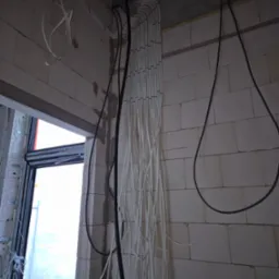 Białe i czarne przewody ułożone pionowo w rurkach na ścianie z białej cegły, widoczne elementy montażowe na suficie i okno w tle.