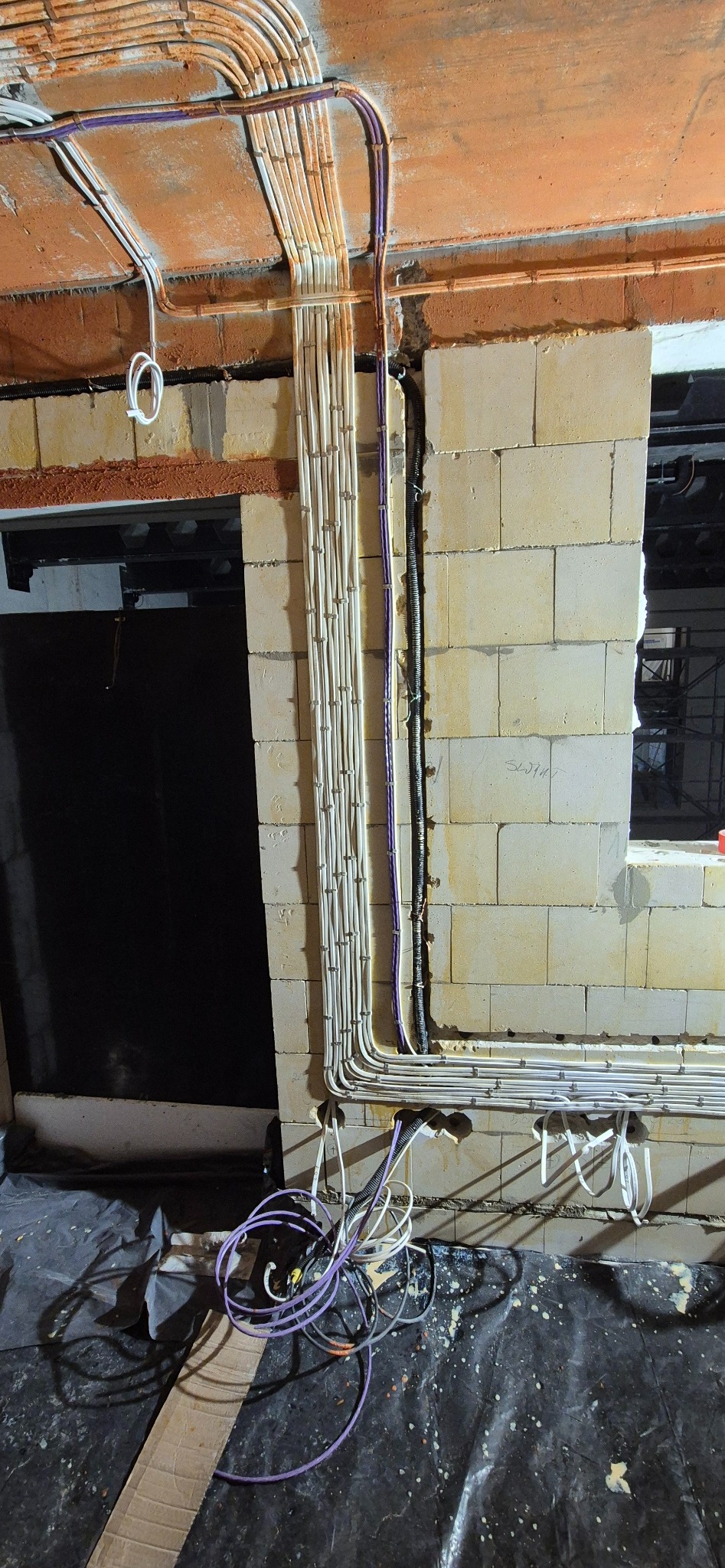 Równolegle ułożone, białe rury instalacyjne na ścianie z pustaków, z widocznymi kablami w kolorze fioletowym i czarnym. Przewody częściowo zwisają na folii ochronnej.