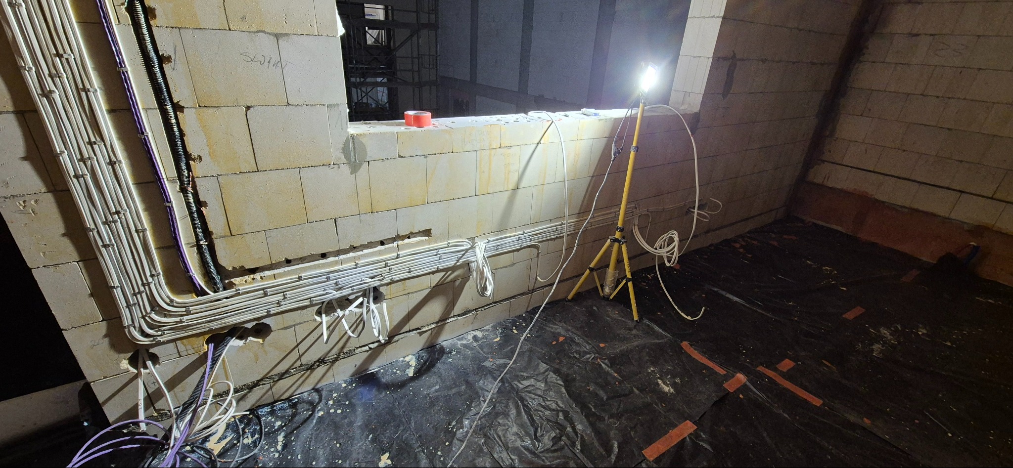 Ściana z instalacją elektryczną w rurkach PCV, zabezpieczona folią podłoga i lampa na statywie. Widoczne kable w kolorach białym, czarnym i fioletowym.