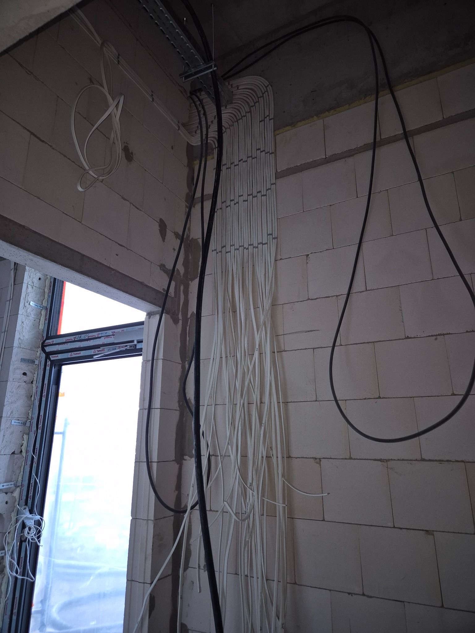 Białe i czarne przewody ułożone pionowo w rurkach na ścianie z białej cegły, widoczne elementy montażowe na suficie i okno w tle.