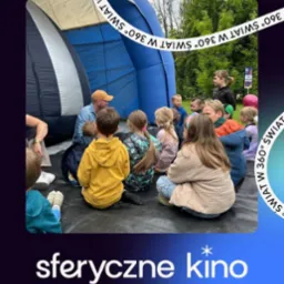 Grupa dzieci siedzi na podłodze, słuchając mężczyzny w pomarańczowej czapce przed wejściem do sferycznego kina. W tle zieleń drzew.