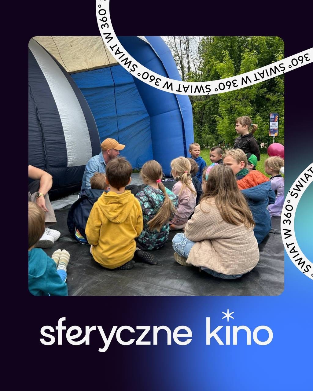 Grupa dzieci siedzi na podłodze, słuchając mężczyzny w pomarańczowej czapce przed wejściem do sferycznego kina. W tle zieleń drzew.