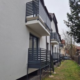 TMM SPAW YURII BUDZAN - Biały budynek z balkonami z czarnymi, poziomymi barierkami. Widok z boku, ukazujący kilka balkonów z metalowymi schodami. Tło stanowią drzewa i sąsiednie budynki.