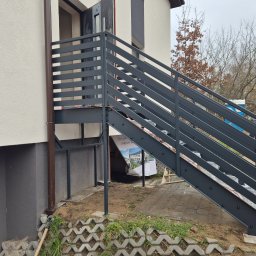 TMM SPAW YURII BUDZAN - Zewnętrzne schody metalowe z szarą, poziomą balustradą, prowadzące do drzwi wejściowych budynku. Widoczne elementy konstrukcyjne i otoczenie w trakcie prac wykończeniowych.