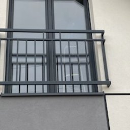 TMM SPAW YURII BUDZAN - Szara, metalowa balustrada okienna z prostych profili, zamontowana na elewacji budynku z oknem w ciemnej ramie. Minimalistyczny design, nowoczesne budownictwo.