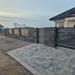 TMM SPAW YURII BUDZAN - Nowoczesne ogrodzenie posesji z przesuwną bramą wjazdową, wykończone szarą kostką brukową. Widoczny fragment domu z ciemnym dachem i jasną elewacją.