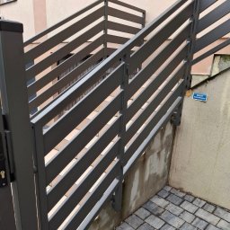 TMM SPAW YURII BUDZAN - Nowoczesna, szara brama metalowa z poziomymi szczeblami, prowadząca do schodów. Widoczny fragment budynku w tle i kostka brukowa na podłożu.