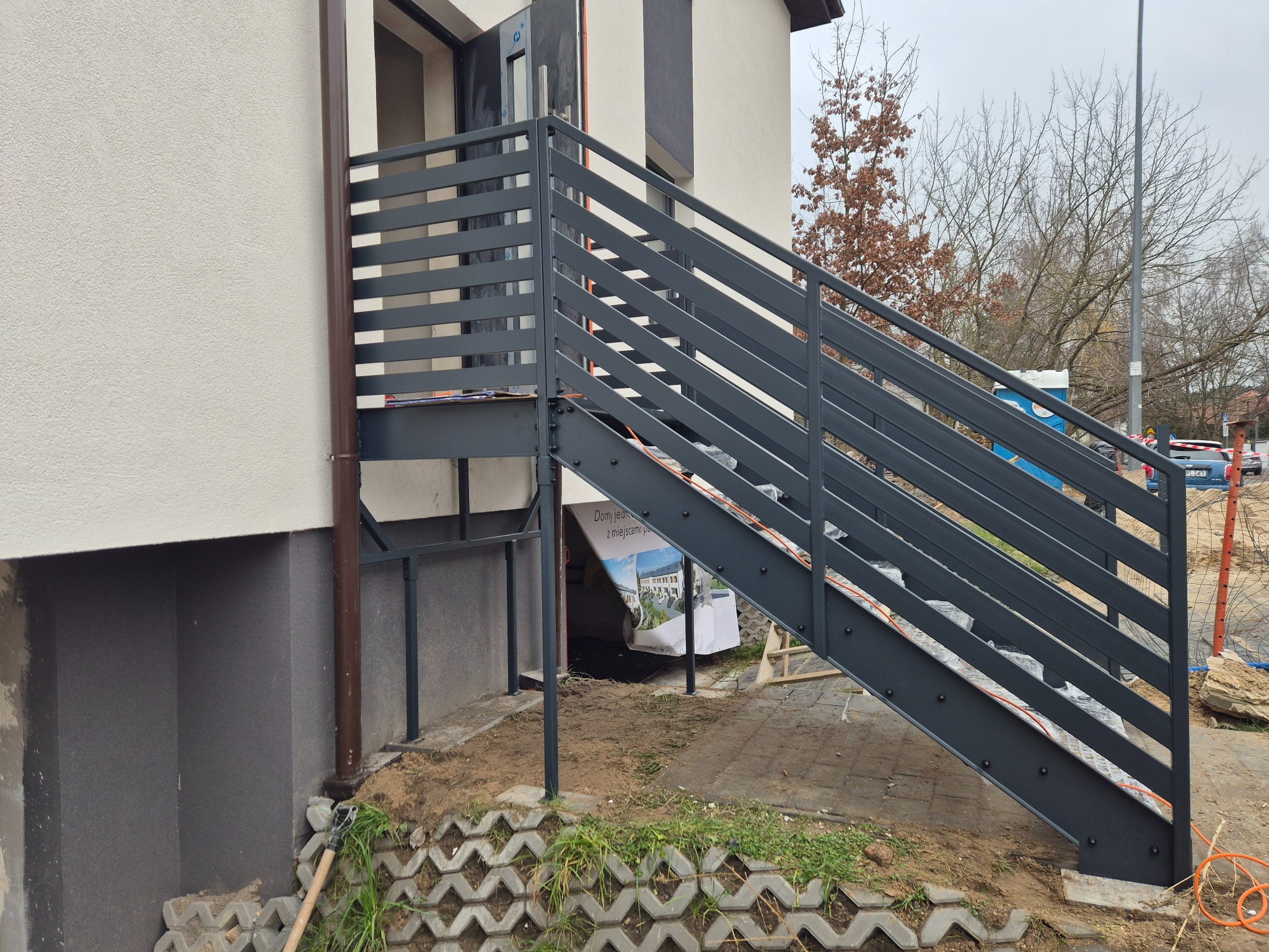 Zewnętrzne schody metalowe z szarą, poziomą balustradą, prowadzące do drzwi wejściowych budynku. Widoczne elementy konstrukcyjne i otoczenie w trakcie prac wykończeniowych.
